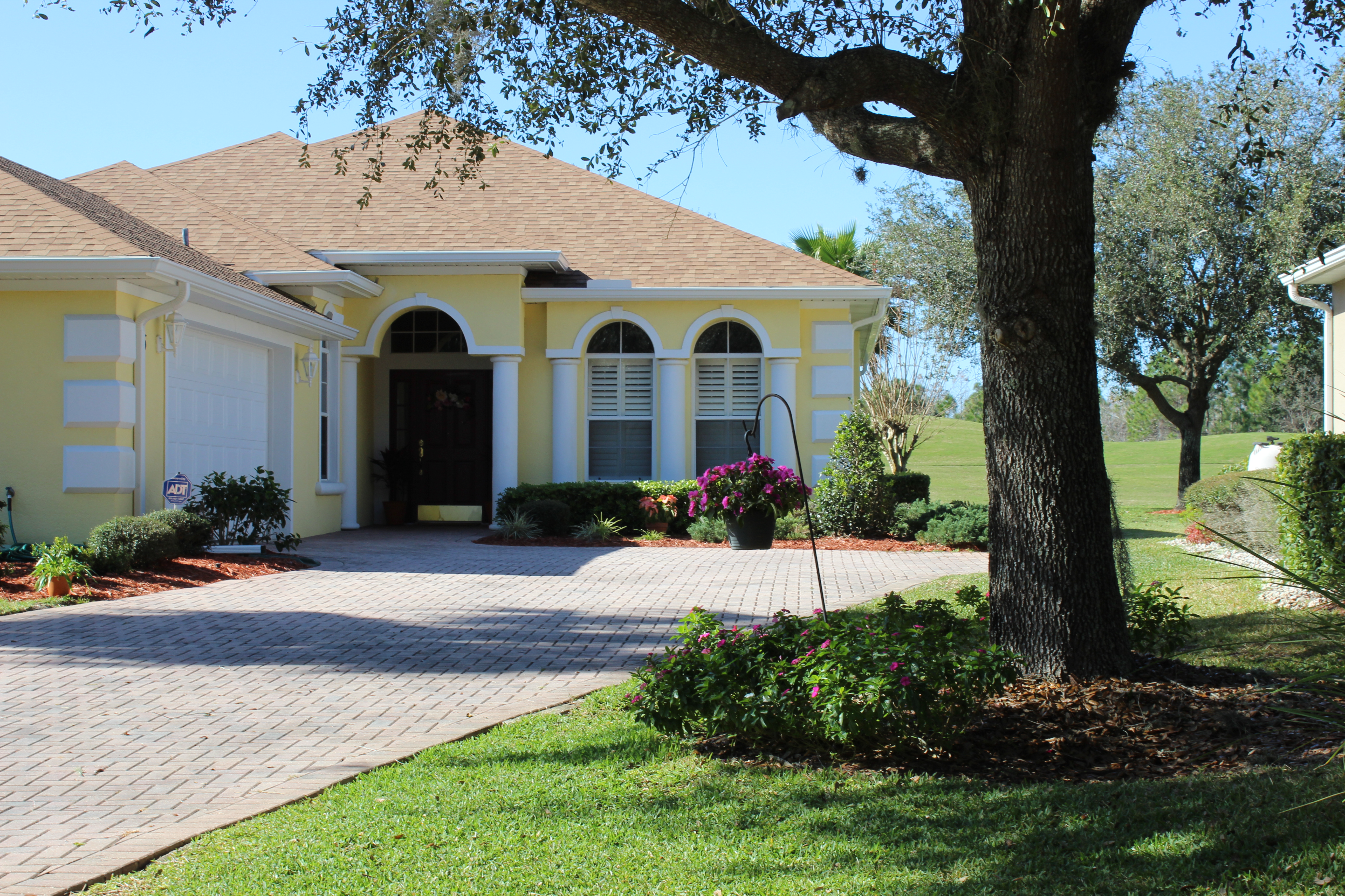 Photograph of yellow house with brick driveway, manicured landscaping and oak tree in yard